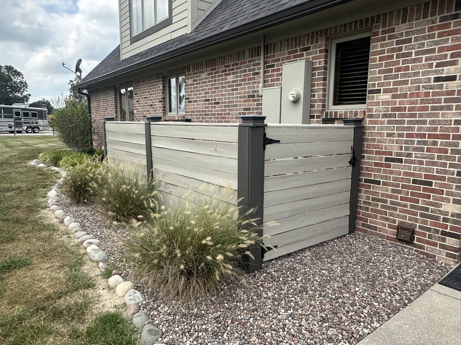 A horizontal slat privacy screen in tan with dark posts, enclosing utilities beside a brick home